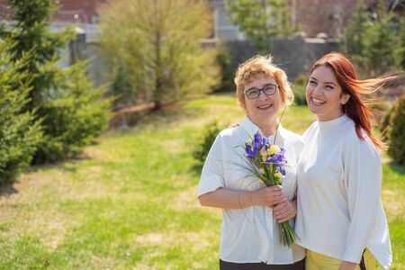 Adult daughter gives flowers to her mother outside, in the courtyard of the house. Spending time...