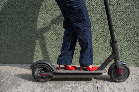 Young woman in formal wear on red hight heels is standing on electrical scooter. Close-up of female...