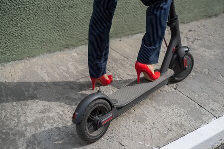 Young woman in formal wear on red hight heels is standing on electrical scooter. Close-up of female...