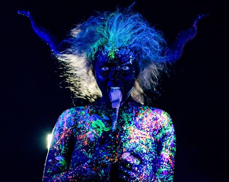 The girl in the image of the devil, contact lenses and horns. Woman paints her tongue in ultraviolet...