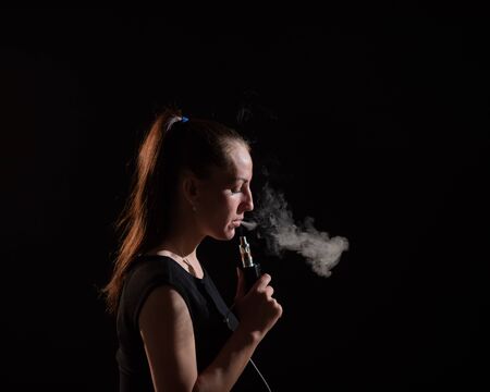 The brunette smokes an electronic cigarette on a black background, releases thick smoke from her...
