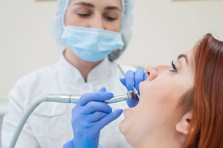 An attractive smiling female doctor in white uniform examines a red-haired female patient. Dentist produces treats teeth with a drill.の写真素材