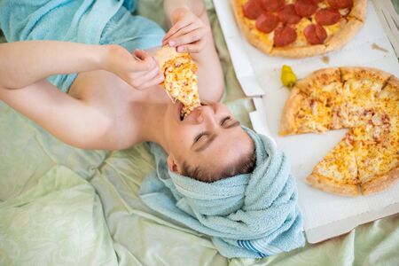 Cute girl with a towel on her head eats pizza in bed. Young woman eating pizza in bed. Life is a pleasure, body positive. Love for Italian food. Food habits, addiction to fast food.の写真素材