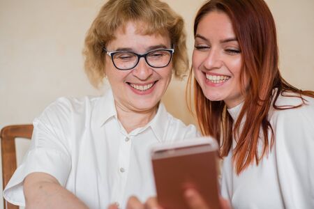 An elderly mother and daughter dressed in light clothes are smiling and taking selfies. Mom and...