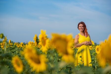 A red-haired woman in a yellow dress is standing in a field of sunflowers. Beautiful girl in a skirt sun enjoys a cloudless day in the countryside. Pink locks of hair.の写真素材
