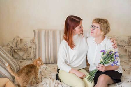 Daughter gives flowers to an adult mother sitting on the sofa in the living room. Spending time together, celebrating at home on weekends. Mothers Day. Warm intergenerational relationshipsの写真素材