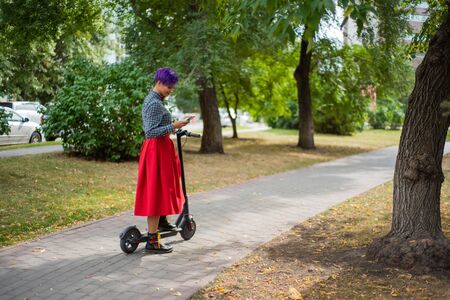 A young woman with purple hair is riding an electric scooter in the park. Stylish girl with a shaven temple in a checkered shirt, a long red skirt and bow-tie writes a message on a smartphone. Hipsterの写真素材