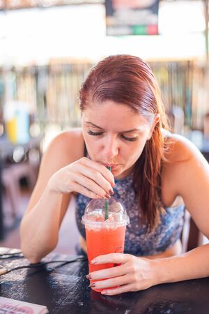 Portrait of a beautiful and cheerful redhead woman with wet hair drinking a refreshing cocktail in a street cafe. Girl on vacation drinks a cocktail after swimmingの写真素材