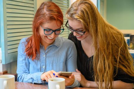 Two excited young girls using mobile phones, sitting in a cafe and pointing finger. Red-haired woman...