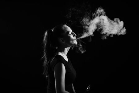 The brunette smokes an electronic cigarette on a black background, releases thick smoke from her...