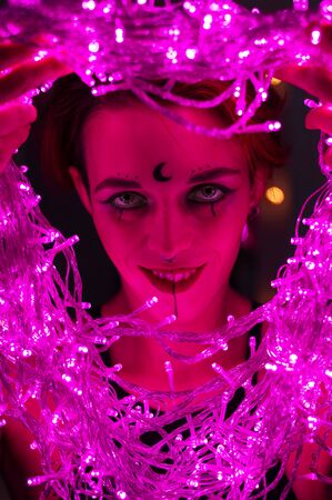 Woman with witch makeup and an earring in her nose. The girl holds a garland of flickering pink lights near her face. Light bulbs illuminate the witchs face in the dark.の写真素材