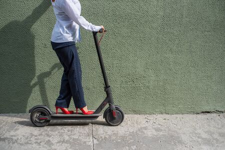 Young woman in formal wear on red hight heels is standing on electrical scooter. Close-up of female legs. A business woman in a trouser suit and red shoes moves around the city on an electric scooterの写真素材