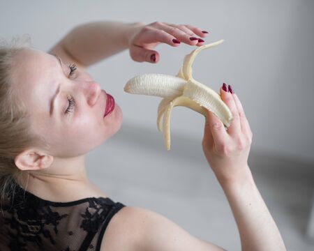 The blonde in black lace underwear erotically eats a banana. Attractive woman with sensual red lips sexually sucks and licks a banana. Oral pleasure.の写真素材