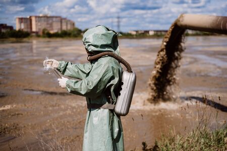 A male environmentalist in a protective suit and gas mask takes a sample of water. Scientist doing...