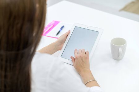 A young woman in a white shirt and jeans sits at a table with a tablet in her hands. A female office...