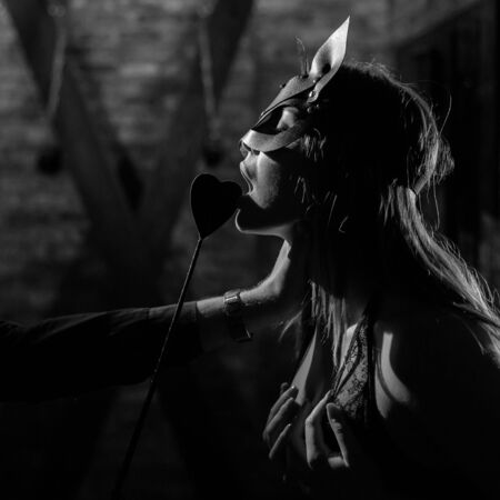 A man in a shirt holds a submissive lover by the neck. A woman in a leather cat mask bites a leather...