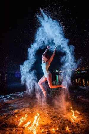 Red-haired woman in a blue swimsuit dancing in clouds of flour in front of a fire. Girl jumping scattering white powder next to a burning flame.の写真素材