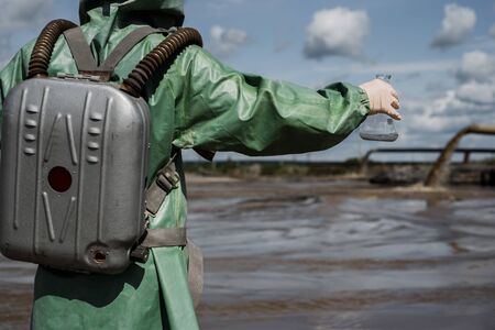 A male environmentalist in a green protective suit and gas mask takes a sample of water in a...