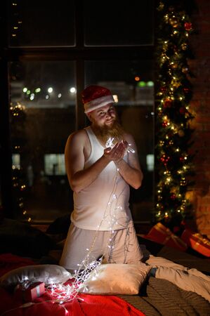 A comic image of santa claus in underwear on christmas eve. A man in a suit of Santa Claus holds a garland with lights in the New Year. A parody of a female photo shoot.の写真素材