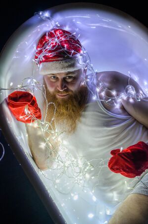 Humorous image of an evil santa claus taking a bath with christmas lights. A man with a red beard in...