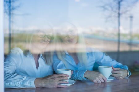 Two friends are enjoying coffee in a coffee shop, looking through a glass with reflections, sitting...