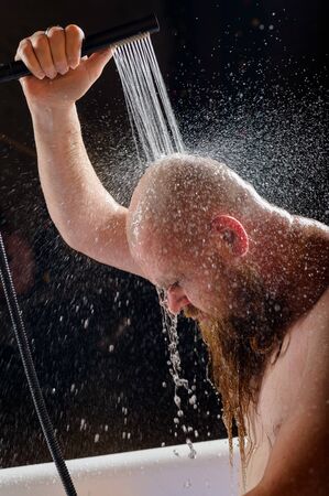 Male takes a shower. A brutal man with a red beard is standing in the bathroom under running water and washes. Spray scatter on a black background.の写真素材