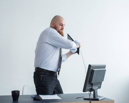Angry bald man with a red beard in the office in a business suit crashes a computer. Manager with a...