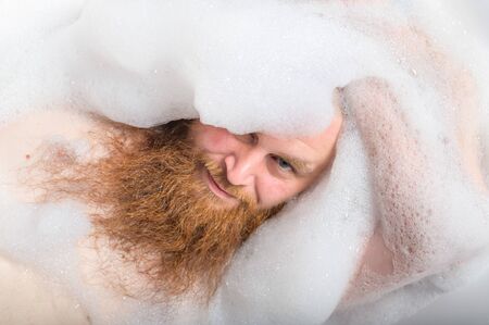 Portrait of a bald man with a long red beard takes a bath with foam. Top view on a cheerful funny...