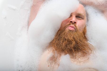 Portrait of a bald man with a long red beard takes a bath with foam. Top view on a cheerful funny guy in soapy water.の写真素材