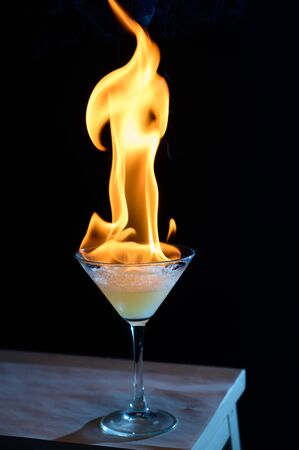 Burning alcoholic cocktail in a martini glass on a table on a black background. Fire drink.