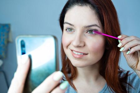 Beautiful redhead woman paints eyelashes and takes a selfie. The girl puts mascara and looks at the...
