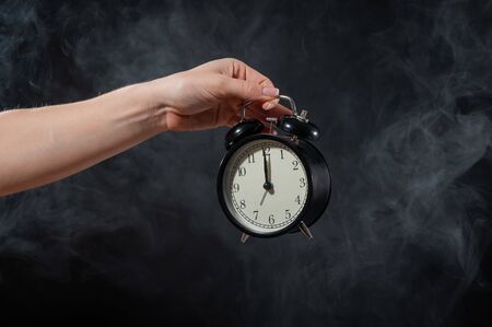 Close-up of a female hand holding a clock on a black background in smoke. Alarm clock at midnight in a mystical fogの写真素材