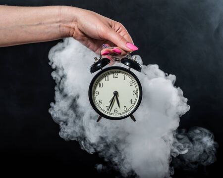 A woman holds an alarm clock in a studio full of smoke. White fog enveloped a round retro mechanical...