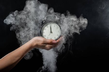Close-up of a female hand holding a clock on a black background in smoke. Alarm clock at midnight in a mystical fog.の写真素材