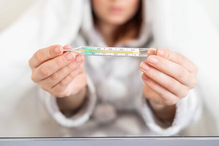 A woman in pajamas with a fever shows a mercury thermometer at the camera. Faceless blurry girl with fever is sitting on the bed.の写真素材