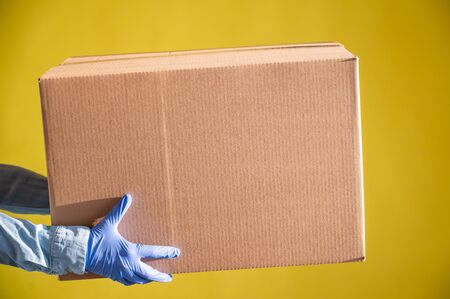 Closeup of female hands in gloves and a denim shirt. The delivery man passes a cardboard box to the customer on a yellow background. Antimicrobial protection in quarantine. Cropped.の写真素材