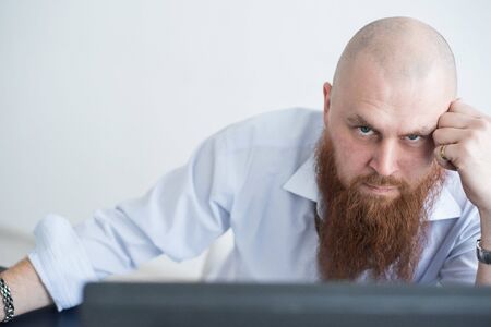 A focused bald man with a red beard stares intently at the camera. Male manager in a white shirt is...