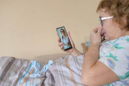 An elderly woman is sick with coronavirus and is in quarantine. A pensioner lies in bed and consults a doctor online about a viral disease.の写真素材