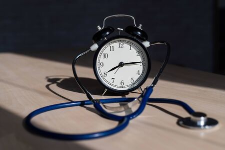 Classic old black mechanical alarm clock with stethoscope on a wooden table. Time to check the heart. Table clock and a device for measuring blood pressure.の写真素材