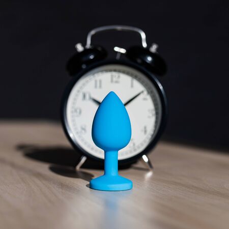 Blue latex anal plug and alarm clock on a wooden table. The time of carnal pleasures.