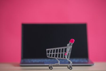 Online shopping concept. Mini grocery cart and laptop on a pink background. Small cart and personal computerの写真素材