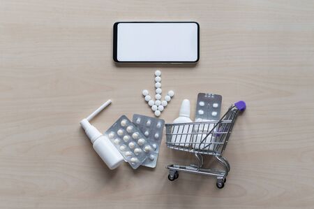 Online pharmacy concept with home delivery. Buying medicine online by phone. Mini trolley with different tablets, sprays, capsules and a smartphone with a white blank screen. Arrow made of pills.の写真素材