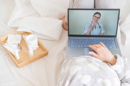 A woman lies on a bed and listens to advice on the treatment of a cold from a therapist. Girl watching a medical transfer on a laptop. The doctor conducts an online consultation.の写真素材