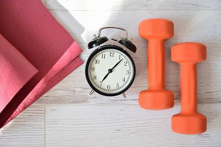 Top view on orange dumbbell alarm clock and raspberry yoga mat. The concept of home sports during...
