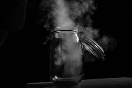 Glass jar with a lid filled with smoke on a black background. Fog in a transparent container on the tableの写真素材