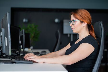 Redhead woman in glasses and casual wear working overtime. Focused girl alone works on a computer in...