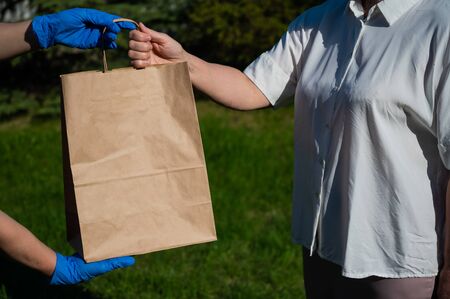 A faceless volunteer with gloves gives an eco friendly paper bag to a retired woman in need. The concept of donation of essential products for the elderly during the coronavirus epidemic.の写真素材