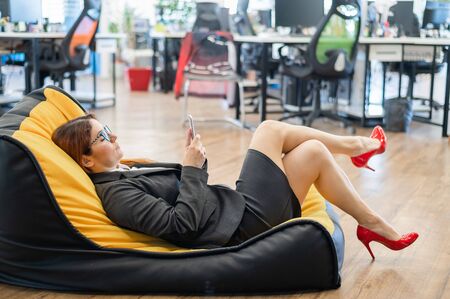 Business woman resting in a cozy bag chair and enjoys mobile. Female Manager at a break in a modern office. Caring for the convenience of employees. Beanbag in the workplace.の写真素材