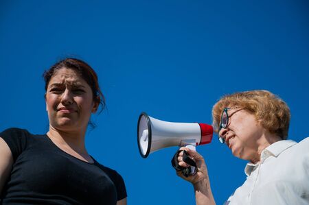 The conflict of generations. An emotional elderly woman shouting at her daughter in a megaphone. An elderly mother swears at a middle-aged woman on a loudspeaker on a blue background.の写真素材