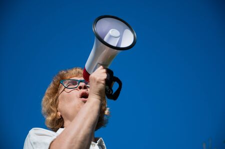 Emotional senior woman makes a speech in a megaphone on the outside. A pensioner yells into a sound amplifier on a blue background. female leader of the rally voices the conditions in the loudspeaker.の写真素材